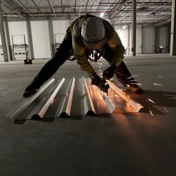 MR montaż Sp. z o.o. - Pracownik w kasku tnie metalową płytę falistą szlifierką kątową, iskry lecą. Hala w tle, Wrocław. Bezpieczeństwo pracy.