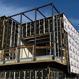 MR montaż Sp. z o.o. - Budynek w trakcie montażu płyt warstwowych z widoczną konstrukcją stalową i izolacją. Ujęcie z dołu, błękitne niebo w tle. Widoczne detale mocowania.