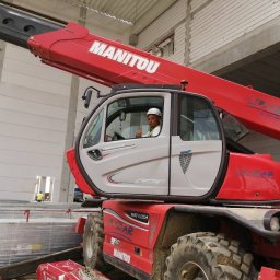 MR montaż Sp. z o.o. - Operator w kabinie czerwonego podnośnika teleskopowego Manitou na budowie hali, z widocznymi płytami warstwowymi i betonową konstrukcją w tle. Kciuk w górę.