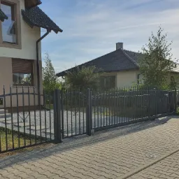 Nowoczesna, przesuwna brama wjazdowa z metalowymi prętami, z dekoracyjnymi elementami w kształcie serc, prowadząca do posesji z kostki brukowej i domu z beżową elewacją.