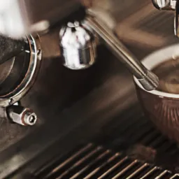 Detaliczny widok na profesjonalny ekspres do kawy podczas przygotowywania espresso do czerwonego kubka, z widoczną dyszą i kroplami kawy wpadającymi do napoju.