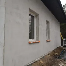 Świeżo otynkowana ściana zewnętrzna budynku z dwoma oknami zabezpieczonymi folią, widoczne ślady po pracach budowlanych na ziemi.