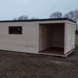 Drewniana konstrukcja altany z oknem, jasne deski, brązowe wykończenia dachu i okna, widoczne deski podłogowe wewnątrz, szare niebo w tle.