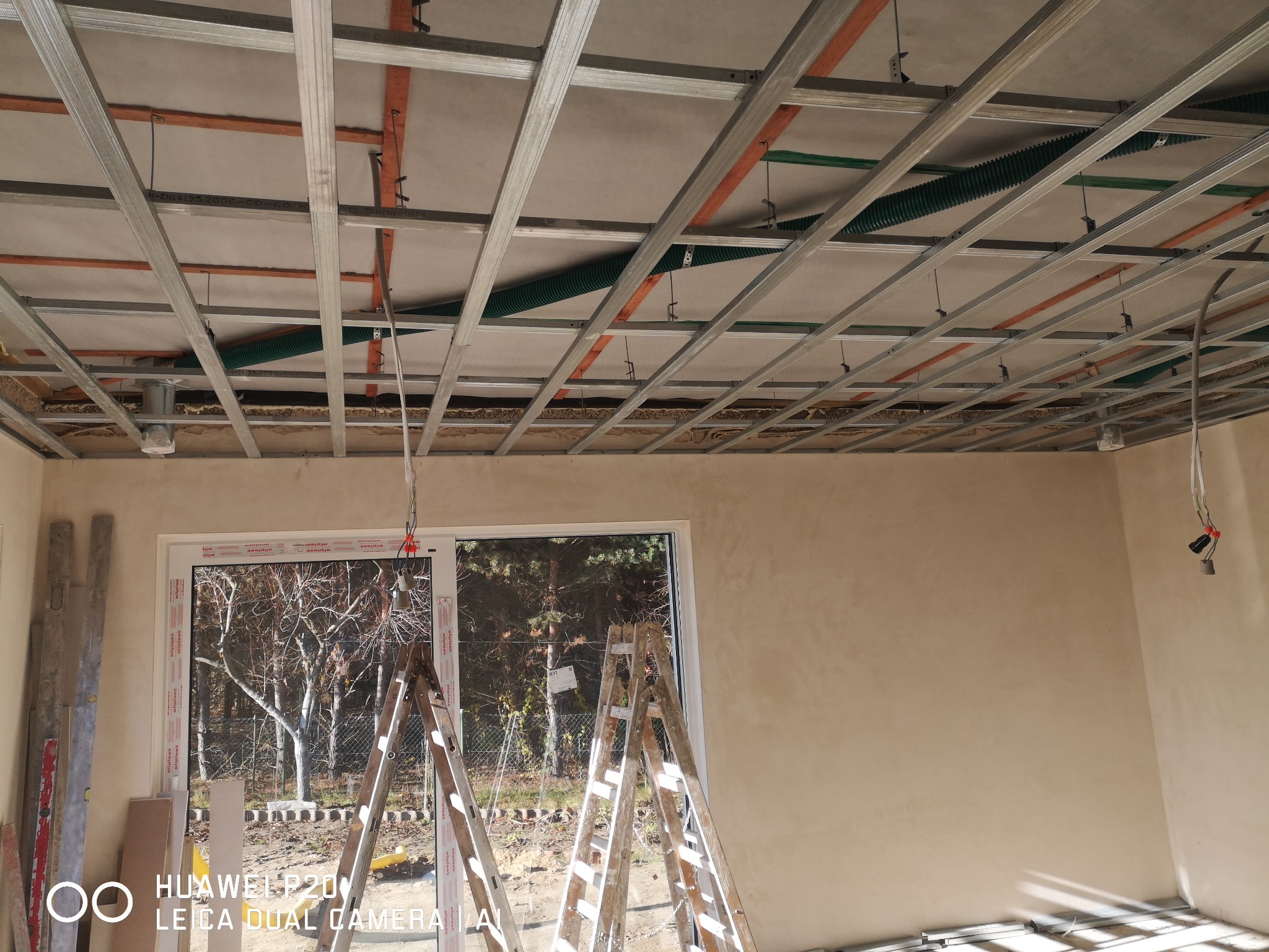 Montaż konstrukcji sufitu podwieszanego z metalowych profili, widoczne przewody wentylacyjne, drabiny opierają się o ścianę z nałożoną warstwą gładzi, okno z widokiem na drzewa w tle.