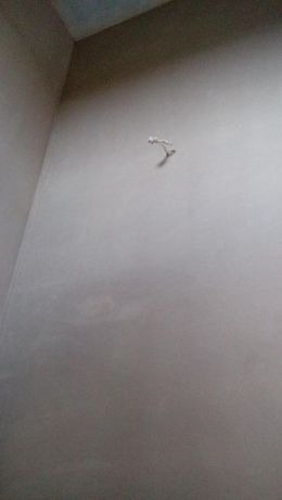 Świeżo otynkowana ściana wewnętrzna z widocznym wystającym przewodem elektrycznym, ukazująca gładką powierzchnię i szare wykończenie.