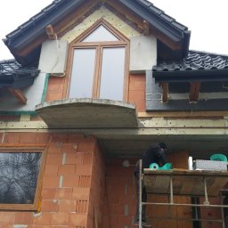 Usługi Ciesielsko-Dekarskie - Budowa domu w trakcie prac: widoczna elewacja z czerwonej cegły, okna z drewnianą ramą, fragment dachu z czarną dachówką oraz rusztowanie z pracownikiem wykonującym prace izolacyjne.