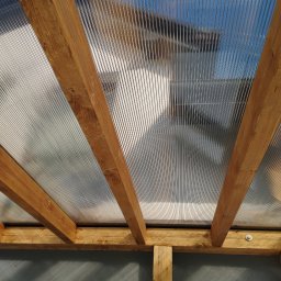 Usługi Ciesielsko-Dekarskie - Widok od dołu na konstrukcję z drewnianych belek i przezroczystego, falistego pokrycia dachowego, przymocowaną śrubami, z widocznym fragmentem ściany w tle.