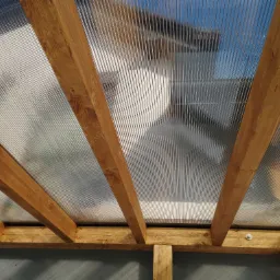 Widok od dołu na konstrukcję z drewnianych belek i przezroczystego, falistego pokrycia dachowego, przymocowaną śrubami, z widocznym fragmentem ściany w tle.