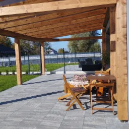 Drewniana pergola z transparentnym zadaszeniem, drewniane meble ogrodowe na szarym brukowanym tarasie, widok na zadbany trawnik i ogrodzenie z siatki.