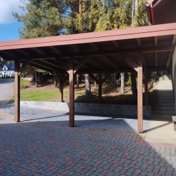 Usługi Ciesielsko-Dekarskie - Drewniana konstrukcja z płaskim dachem, prawdopodobnie zadaszenie miejsca parkingowego, na tle domu i drzew, widok z zewnątrz na kostkę brukową.