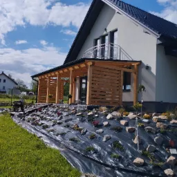 Nowoczesny dom z drewnianą konstrukcją tarasową i przygotowanym podłożem z agrowłókniny, kamieni i roślin ozdobnych.