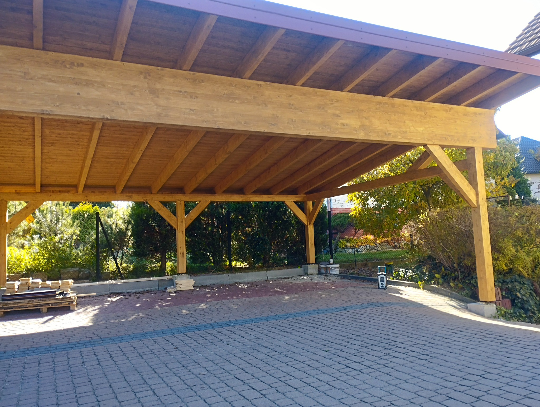 Drewniana konstrukcja zadaszenia nad podjazdem z kostki brukowej, widoczne elementy konstrukcyjne i fragment dachu, w tle zieleń ogrodu.