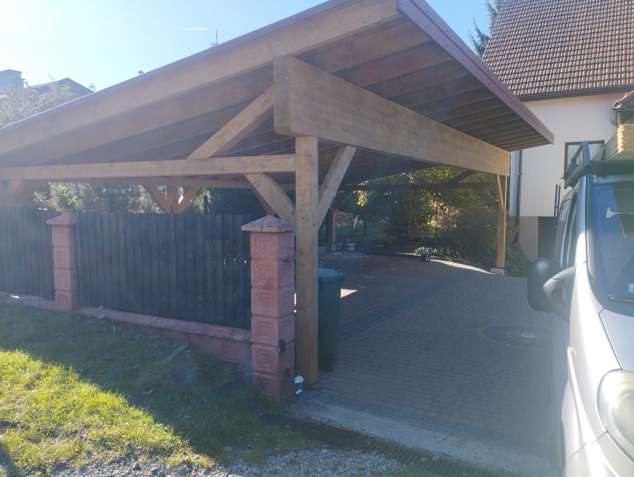 Drewniana konstrukcja zadaszenia nad podjazdem, widoczna z boku, z elementami muru z cegły i zaparkowanym samochodem dostawczym w tle.
