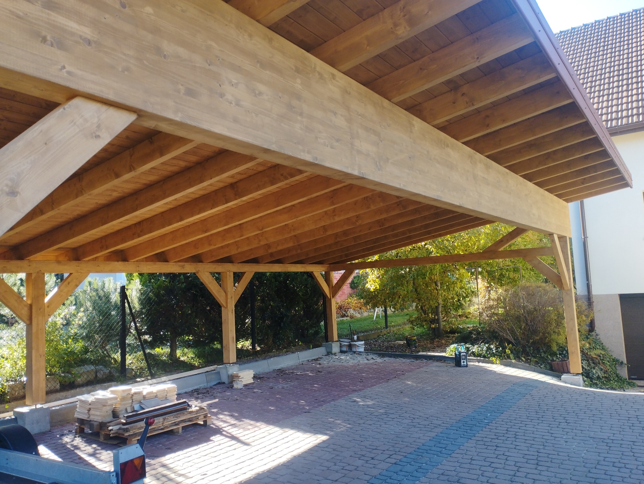 Drewniana konstrukcja z belek i słupów, zadaszenie nad brukowanym podjazdem, widoczne elementy konstrukcyjne i przygotowane materiały budowlane.