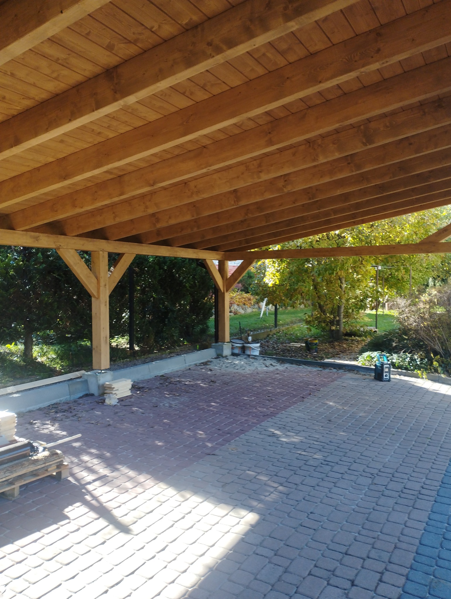 Drewniana konstrukcja z belek i słupów, prawdopodobnie zadaszenie tarasu lub wiaty, widok od dołu, z kostką brukową na ziemi i zielenią w tle.