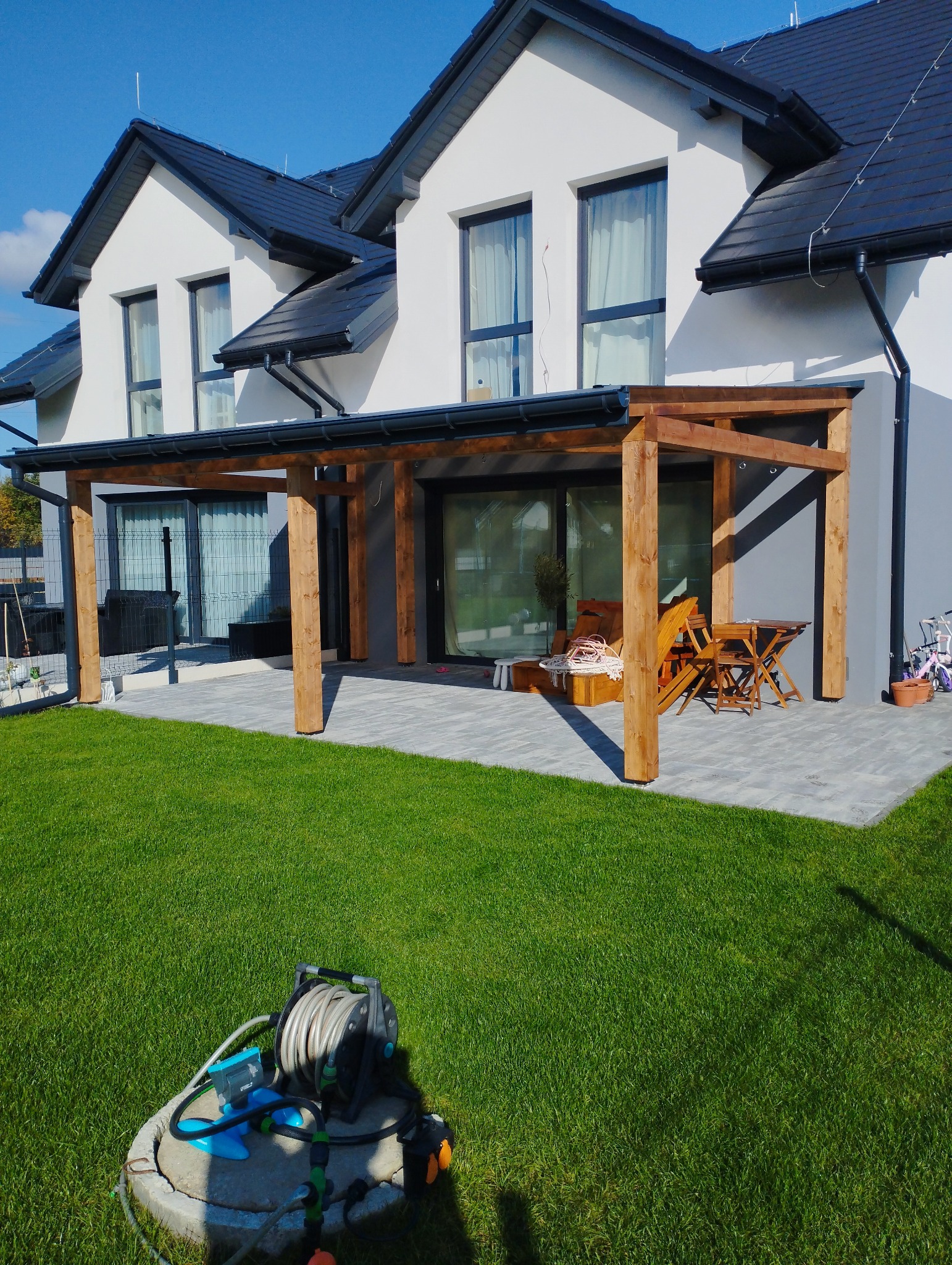 Nowoczesny dom z drewnianą konstrukcją zadaszenia tarasu, widok z trawnika z wężem ogrodowym w pierwszym planie.