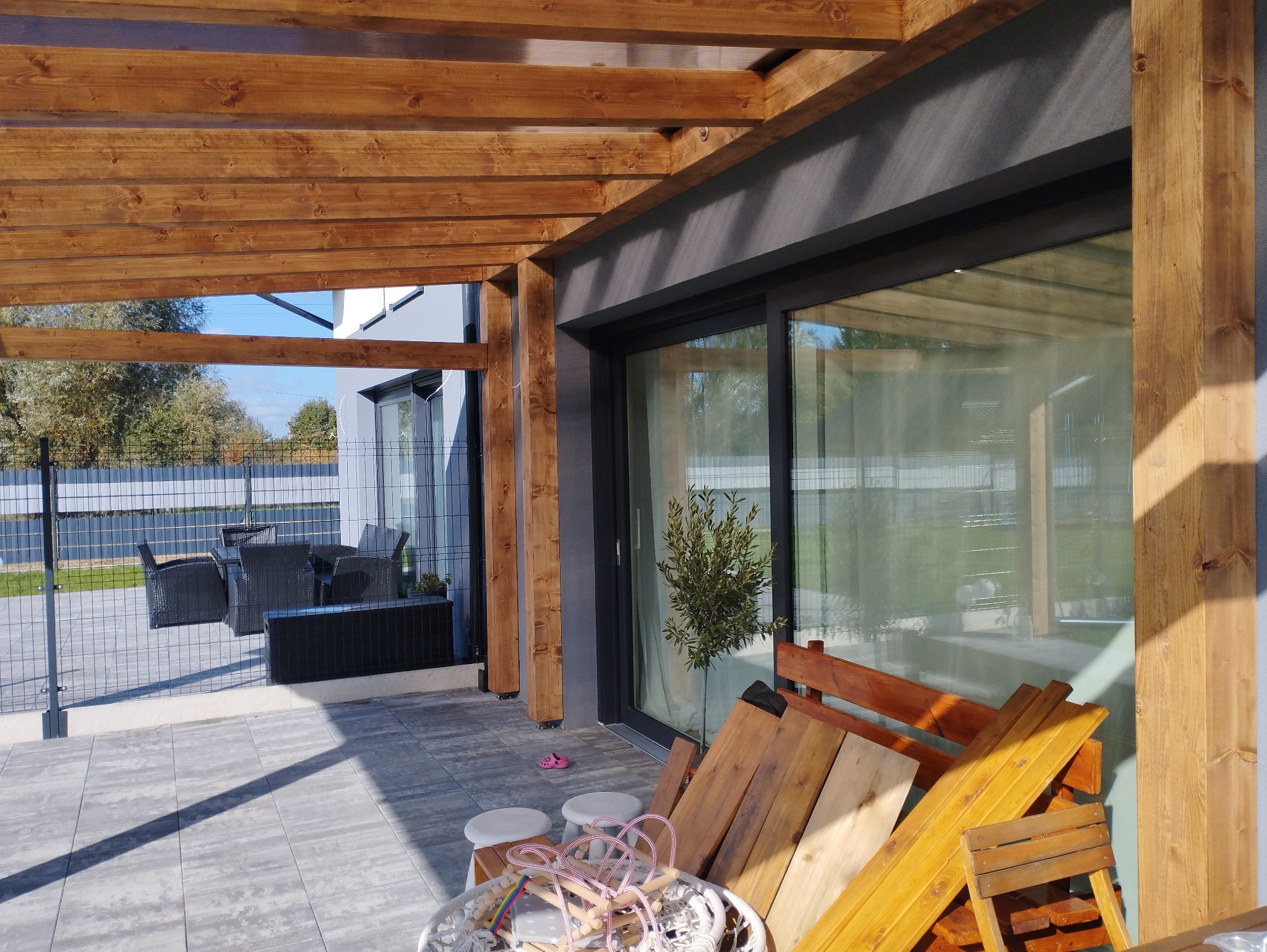 Drewniana konstrukcja z belek nad tarasem, z widocznymi elementami wyposażenia: stolik, krzesła, zabawki i przygotowane deski.