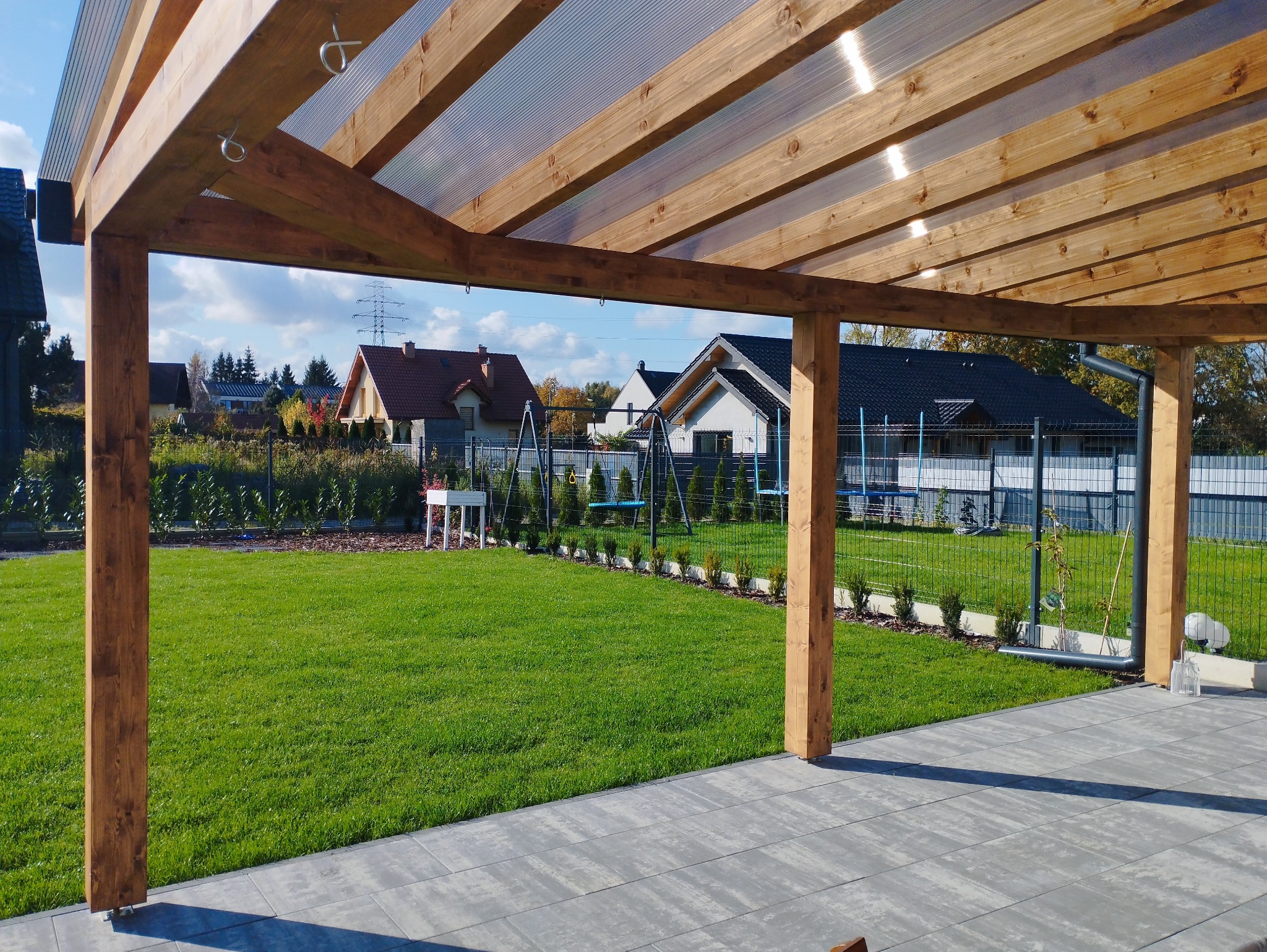 Nowoczesna drewniana konstrukcja z przezroczystym zadaszeniem, widok na zadbany trawnik, huśtawki i zabudowę jednorodzinną w tle.
