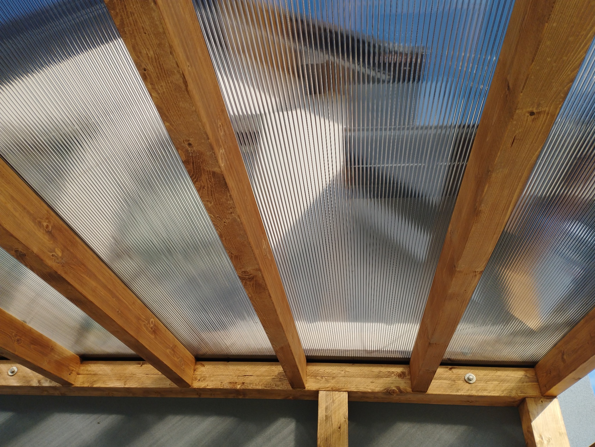 Widok od dołu na konstrukcję z drewnianych belek i przezroczystego, falistego pokrycia dachowego, przymocowaną śrubami, z widocznym fragmentem ściany w tle.