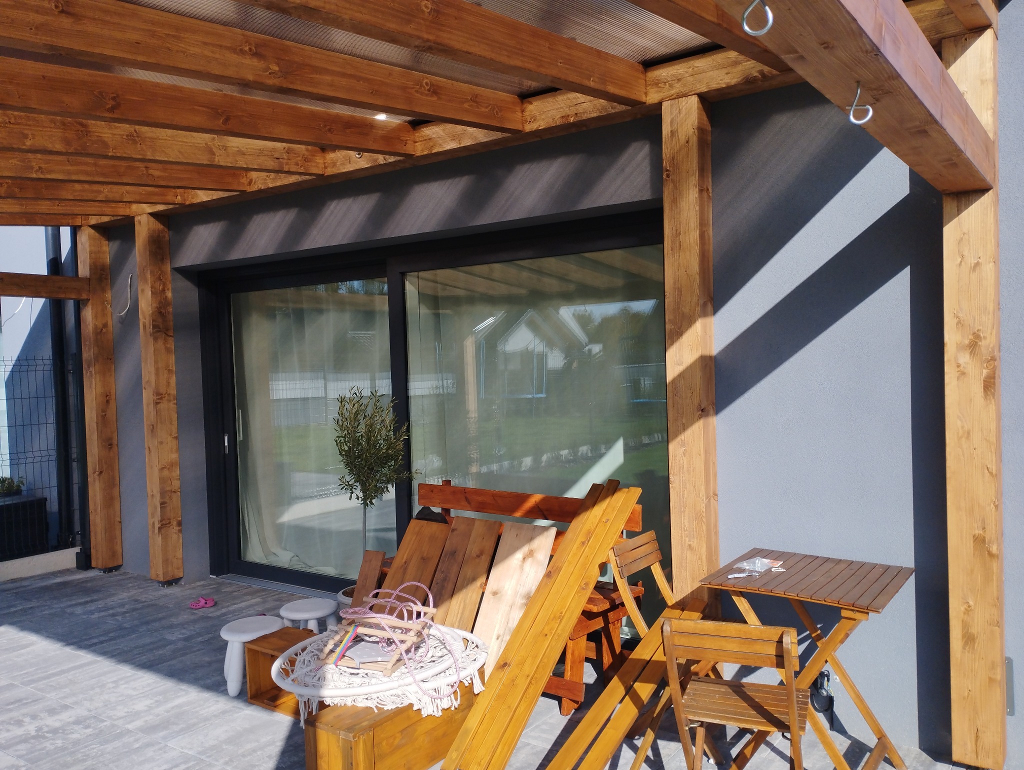 Drewniana konstrukcja z zadaszeniem, prawdopodobnie w trakcie budowy, z widocznymi elementami drewnianymi, metalowymi haczykami i meblami ogrodowymi w tle przesuwne okno domu.