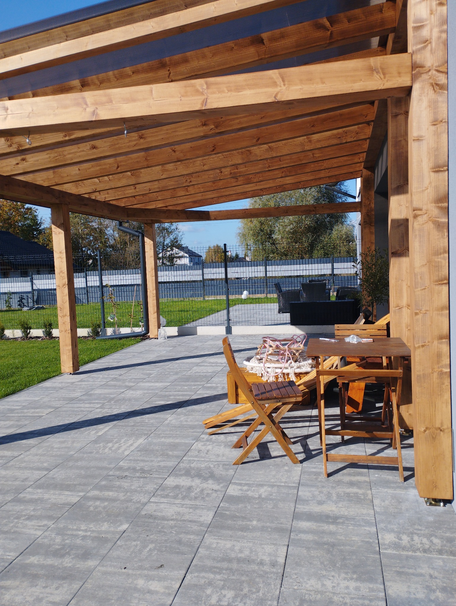Drewniana pergola z transparentnym zadaszeniem, drewniane meble ogrodowe na szarym brukowanym tarasie, widok na zadbany trawnik i ogrodzenie z siatki.