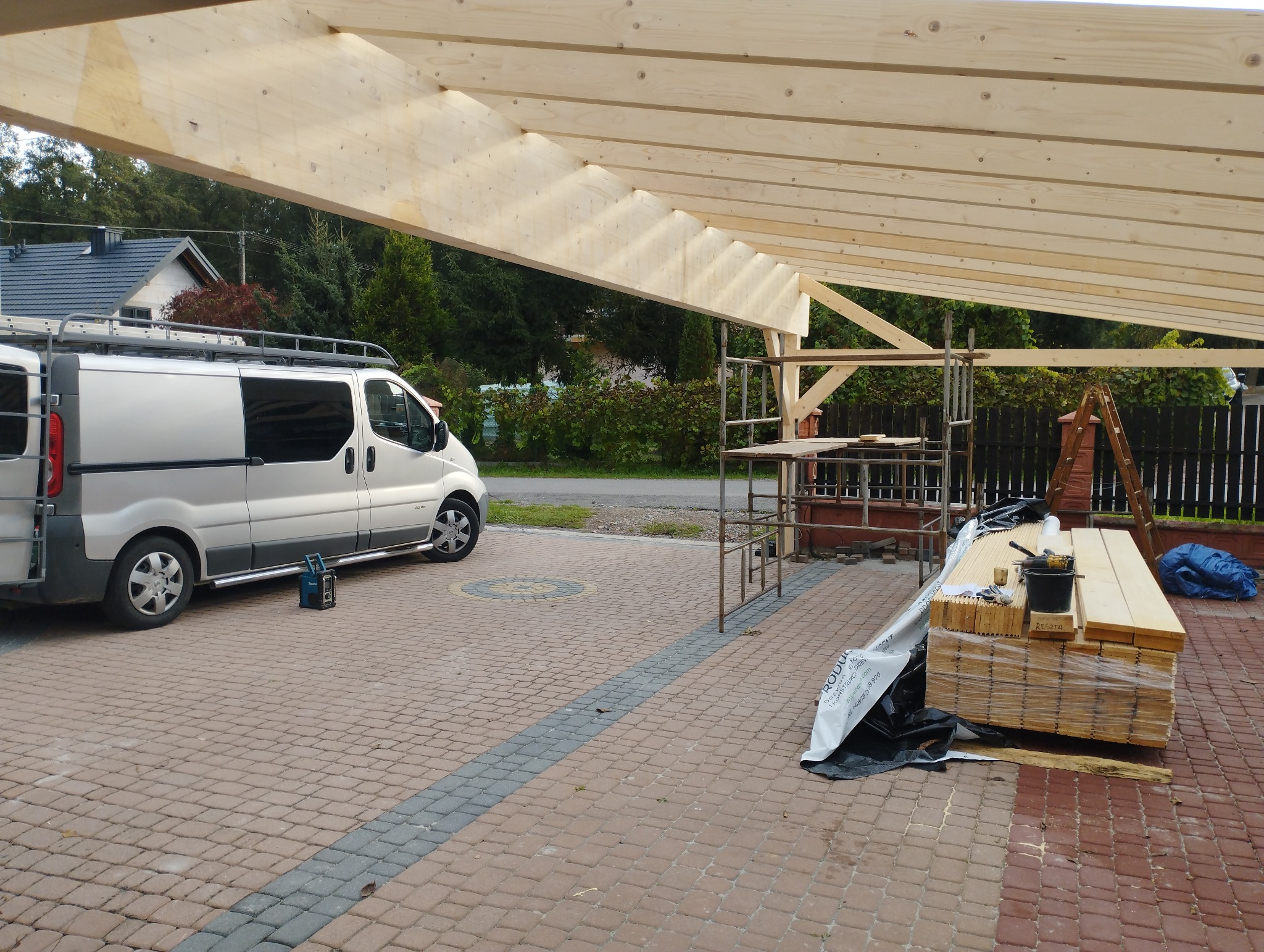 Konstrukcja z belek drewnianych w trakcie montażu, widoczny srebrny bus z drabiną na dachu, rusztowanie, deski i narzędzia na posesji wyłożonej kostką brukową.