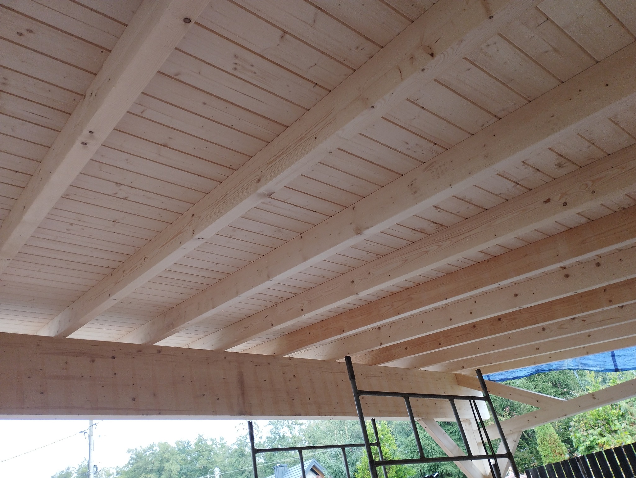 Drewniany strop z belek i desek, widok od dołu, konstrukcja w trakcie montażu, drabina w tle, widok na zewnątrz.