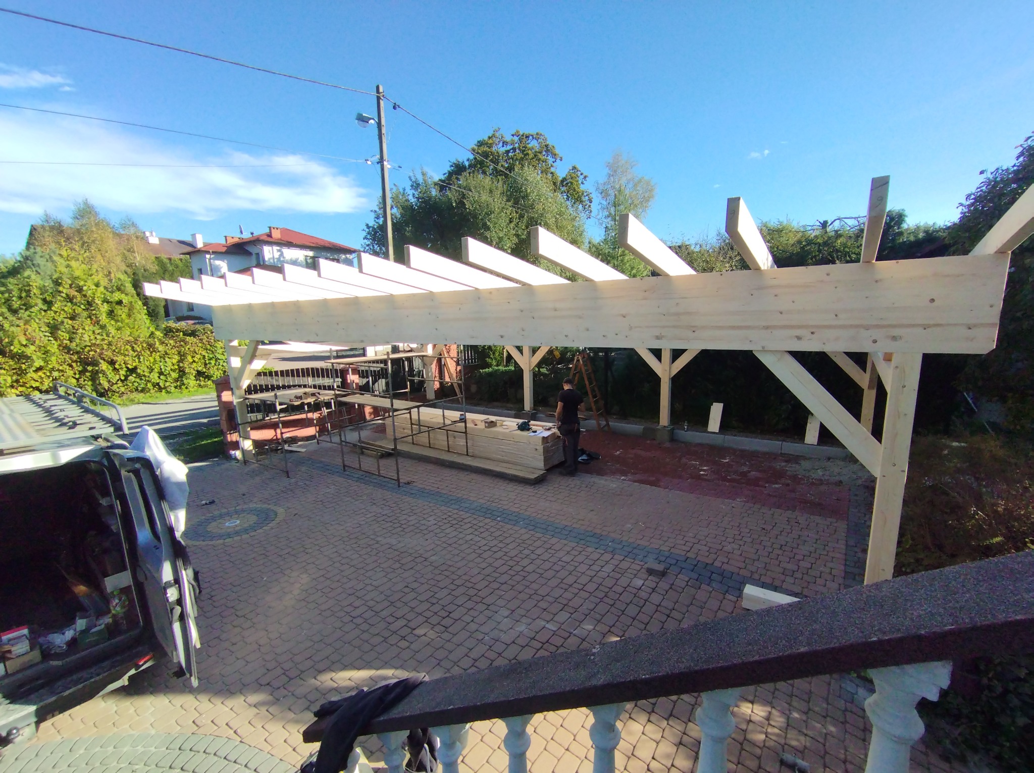 Konstrukcja drewniana w trakcie budowy, prawdopodobnie zadaszenie tarasu lub pergola, widok z góry z balustrady, na pierwszym planie kostka brukowa i zaparkowany samochód dostawczy