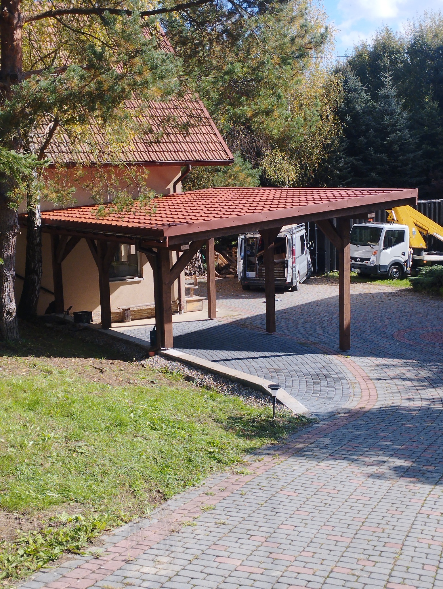 Drewniana konstrukcja z czerwonym dachem, pełniąca funkcję zadaszonego miejsca parkingowego, widoczna na tle domu i podjazdu z kostki brukowej. W tle zaparkowane samochody dostawcze.