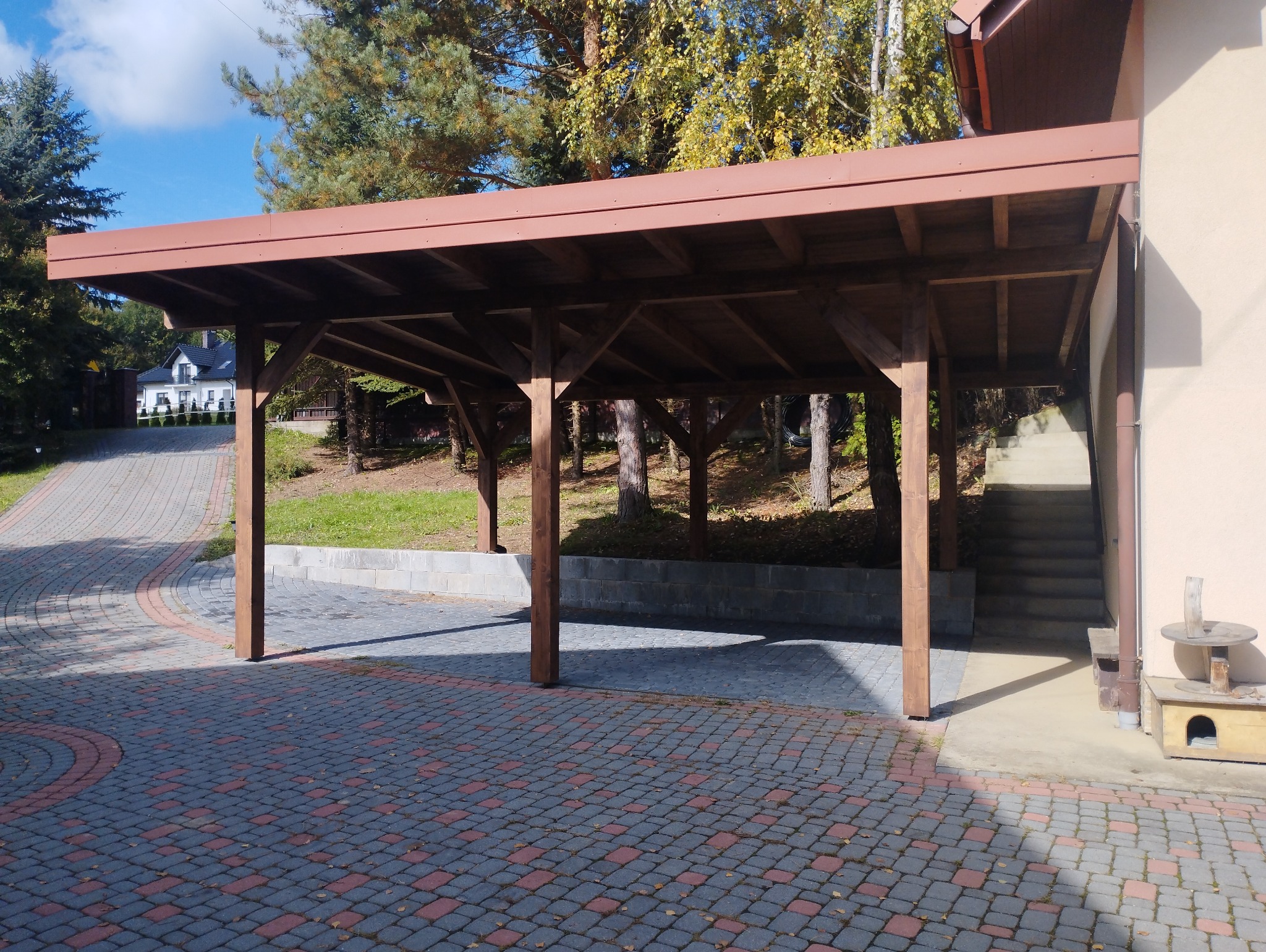 Drewniana konstrukcja z płaskim dachem, prawdopodobnie zadaszenie miejsca parkingowego, na tle domu i drzew, widok z zewnątrz na kostkę brukową.