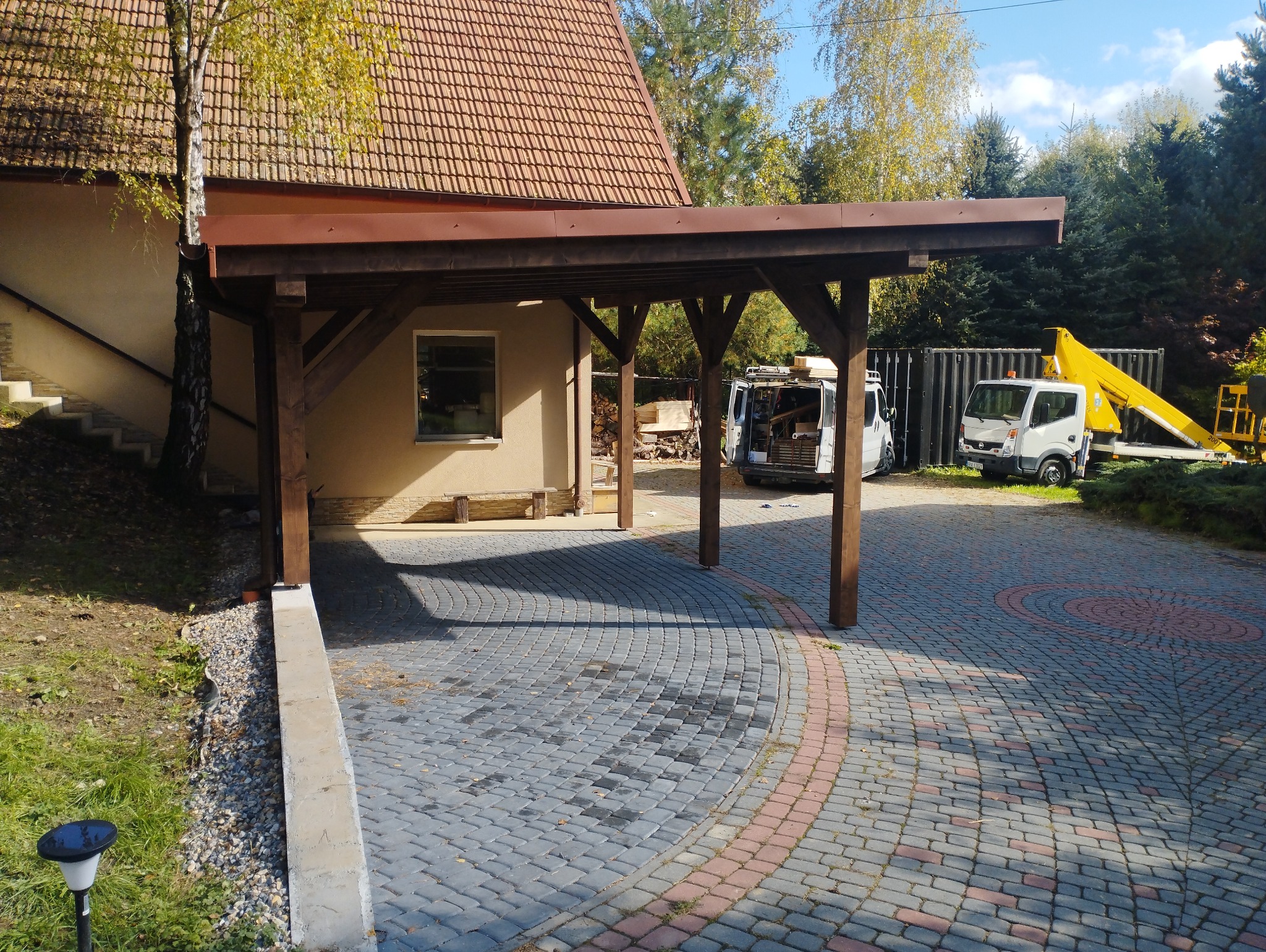 Nowoczesna, drewniana wiata garażowa z brązowym dachem, przylegająca do budynku z jasną elewacją i czerwoną dachówką, na posesji z brukowanym podjazdem i widocznym samochodem dostawczym...