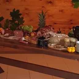 Stół z drewnianym blatem, zastawiony wędlinami, pieczenią, pieczywem, ananasem i innymi przekąskami na tle drewnianej ściany z wiszącymi wędzonkami i dekoracjami roślinnymi.