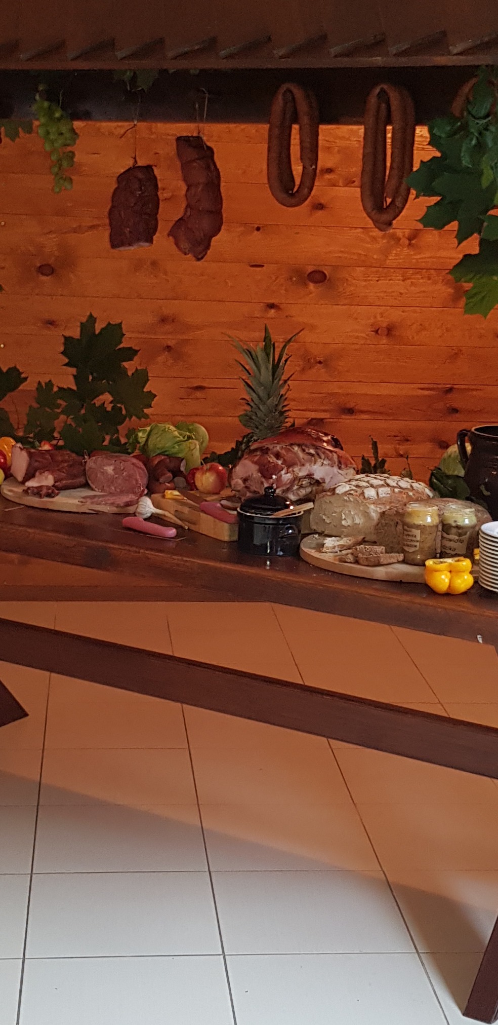 Stół z drewnianym blatem, zastawiony wędlinami, pieczenią, pieczywem, ananasem i innymi przekąskami na tle drewnianej ściany z wiszącymi wędzonkami i dekoracjami roślinnymi.