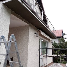 Paweł Pełech - Elewacja domu po renowacji z rusztowaniem i drabiną. Jasna fasada z balkonem i metalową balustradą. Widoczne lampy zewnętrzne i okno.