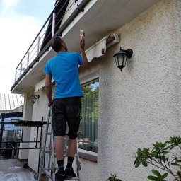 Paweł Pełech - Mężczyzna na drabinie maluje elewację domu pędzlem. Widoczne zabezpieczone okno, lampa zewnętrzna i balkon z metalową barierką. Ujęcie z dołu.