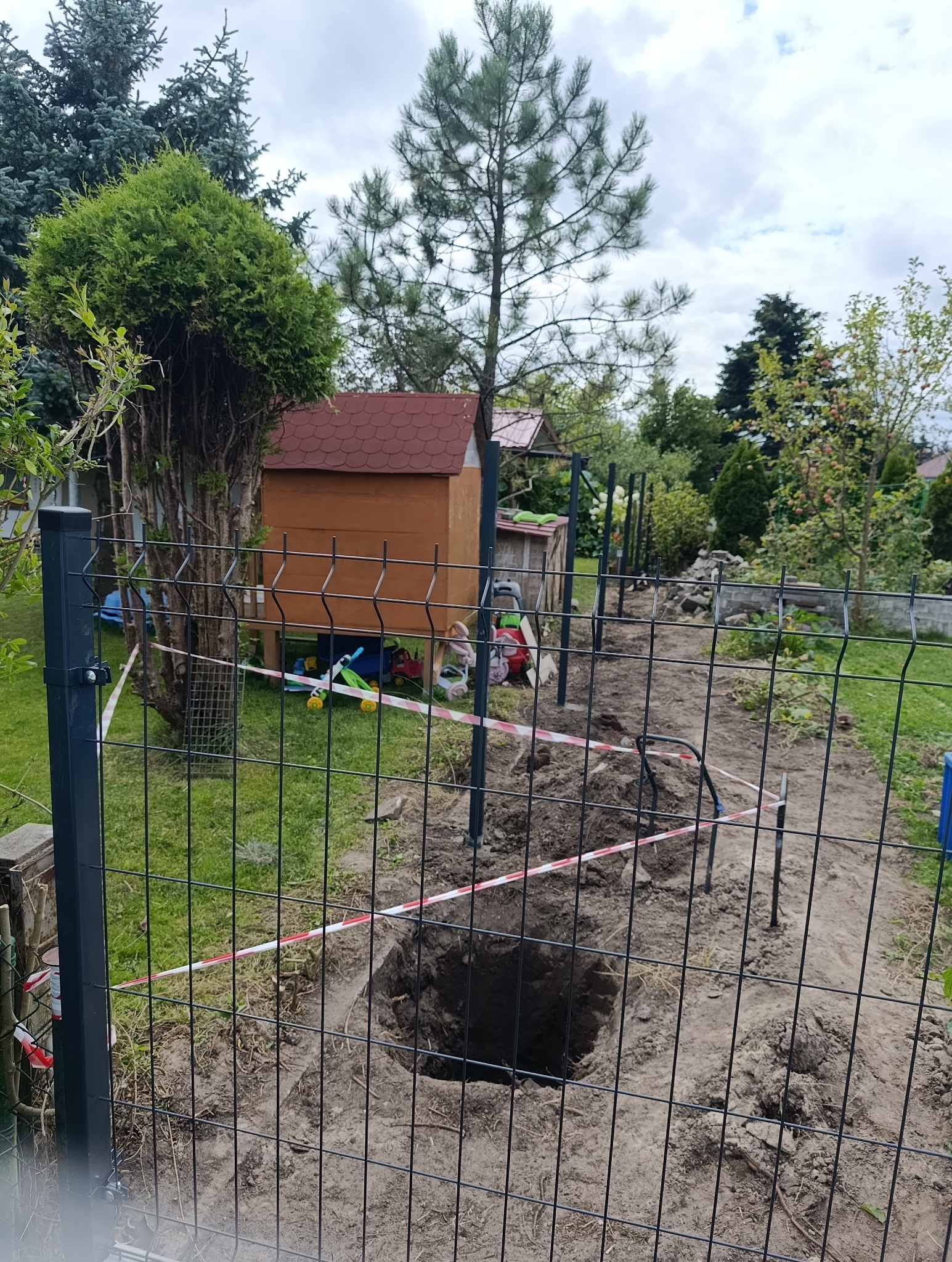 Montaż ogrodzenia panelowego: wykopane dołki pod słupki, teren zabezpieczony taśmą ostrzegawczą. W tle domek narzędziowy i zieleń ogrodu.