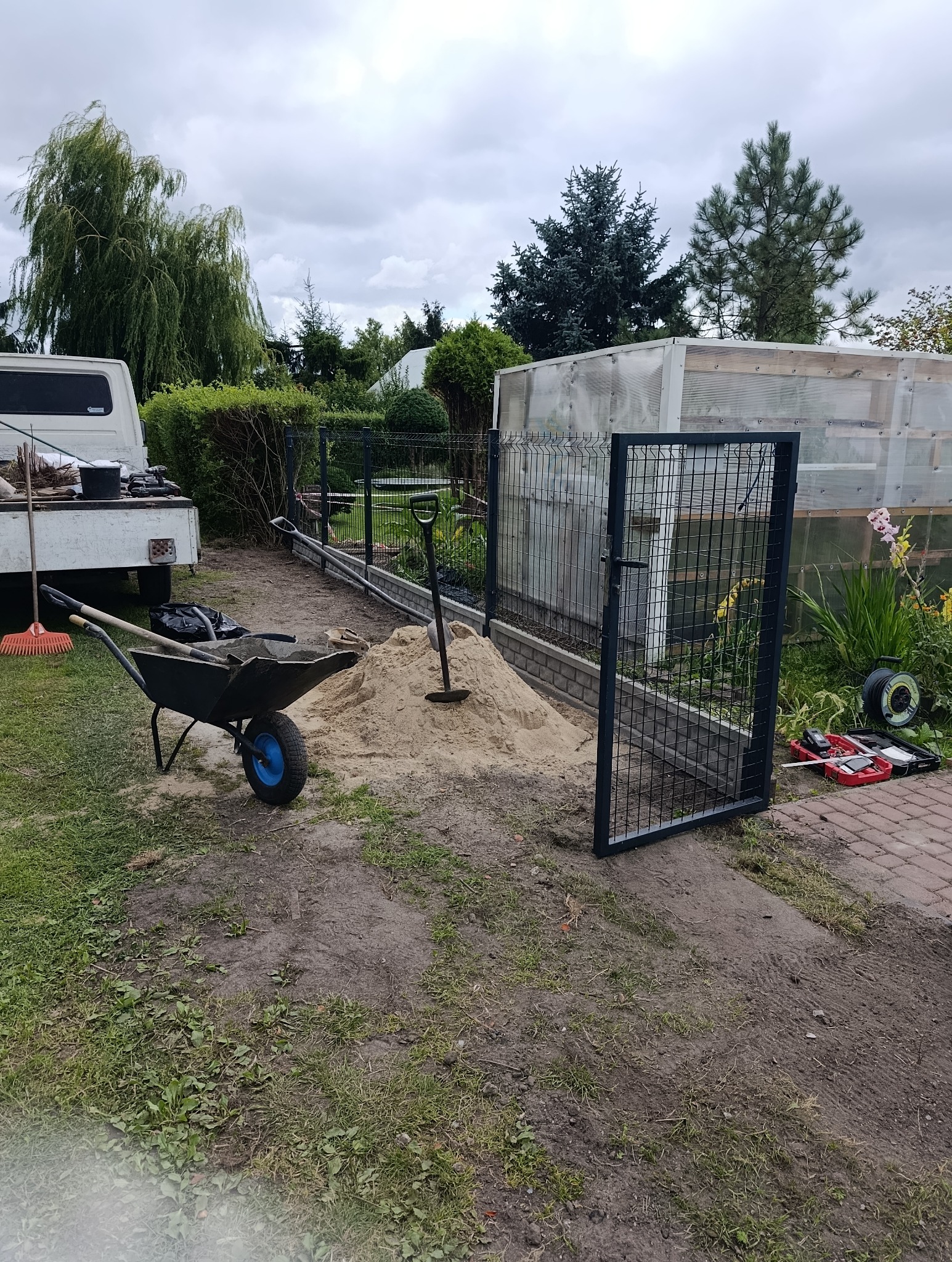 Montaż ogrodzenia panelowego: brama otwarta, widoczny fundament z bloczków, narzędzia, taczka z piaskiem, przygotowany teren. W tle szklarnia i zieleń ogrodu.