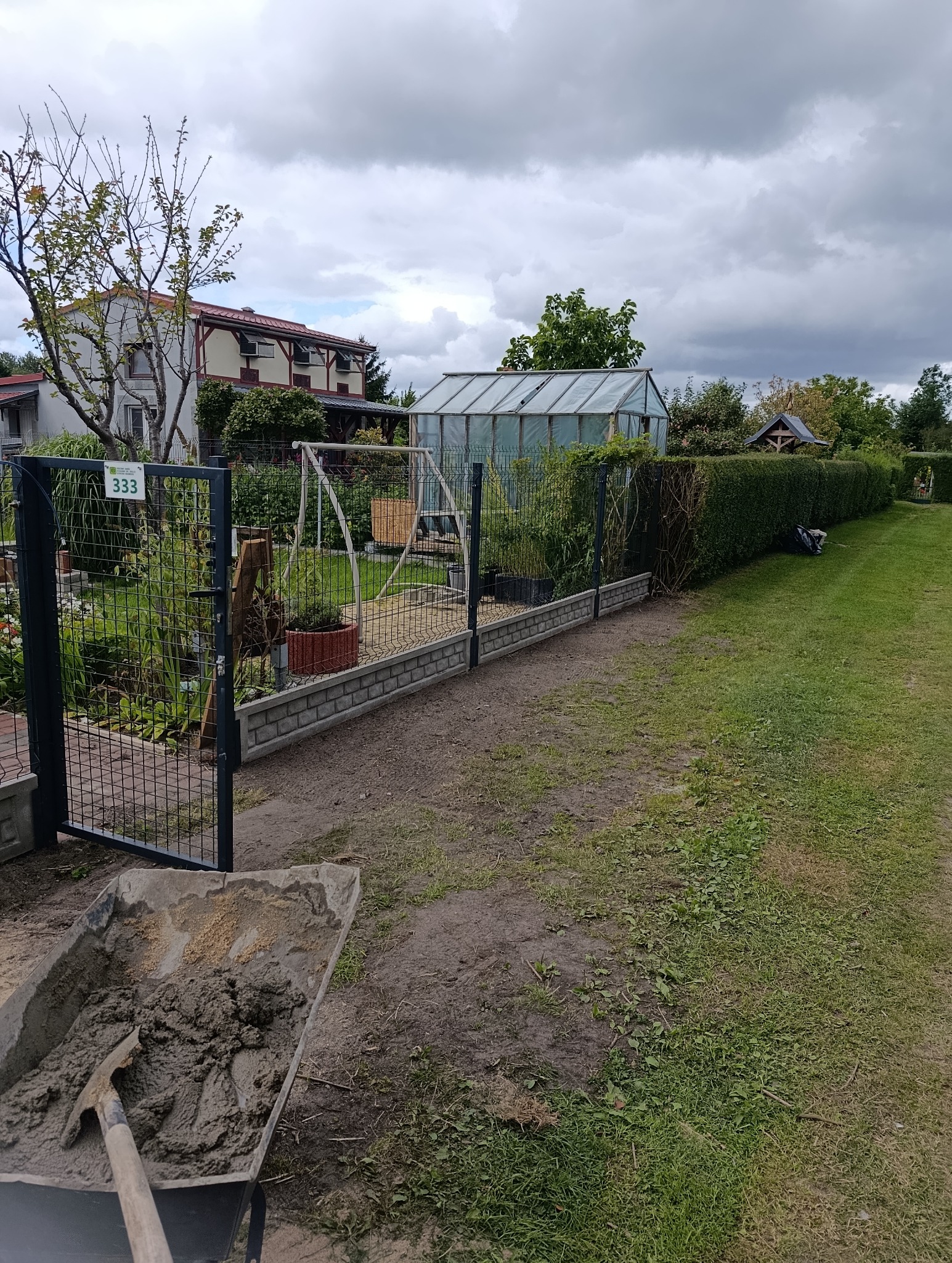 Ogród z nowym ogrodzeniem z prefabrykatów betonowych, widok z wózkiem z zaprawą. W tle szklarnia i dom. Numer 333 na furtce.