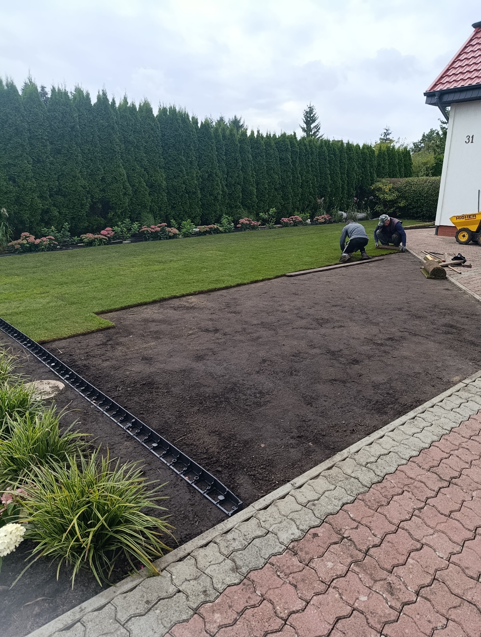 Układanie trawy z rolki na przygotowanym podłożu. Widoczni dwaj pracownicy podczas pracy, częściowo ułożona trawa, system odwadniający i narzędzia.