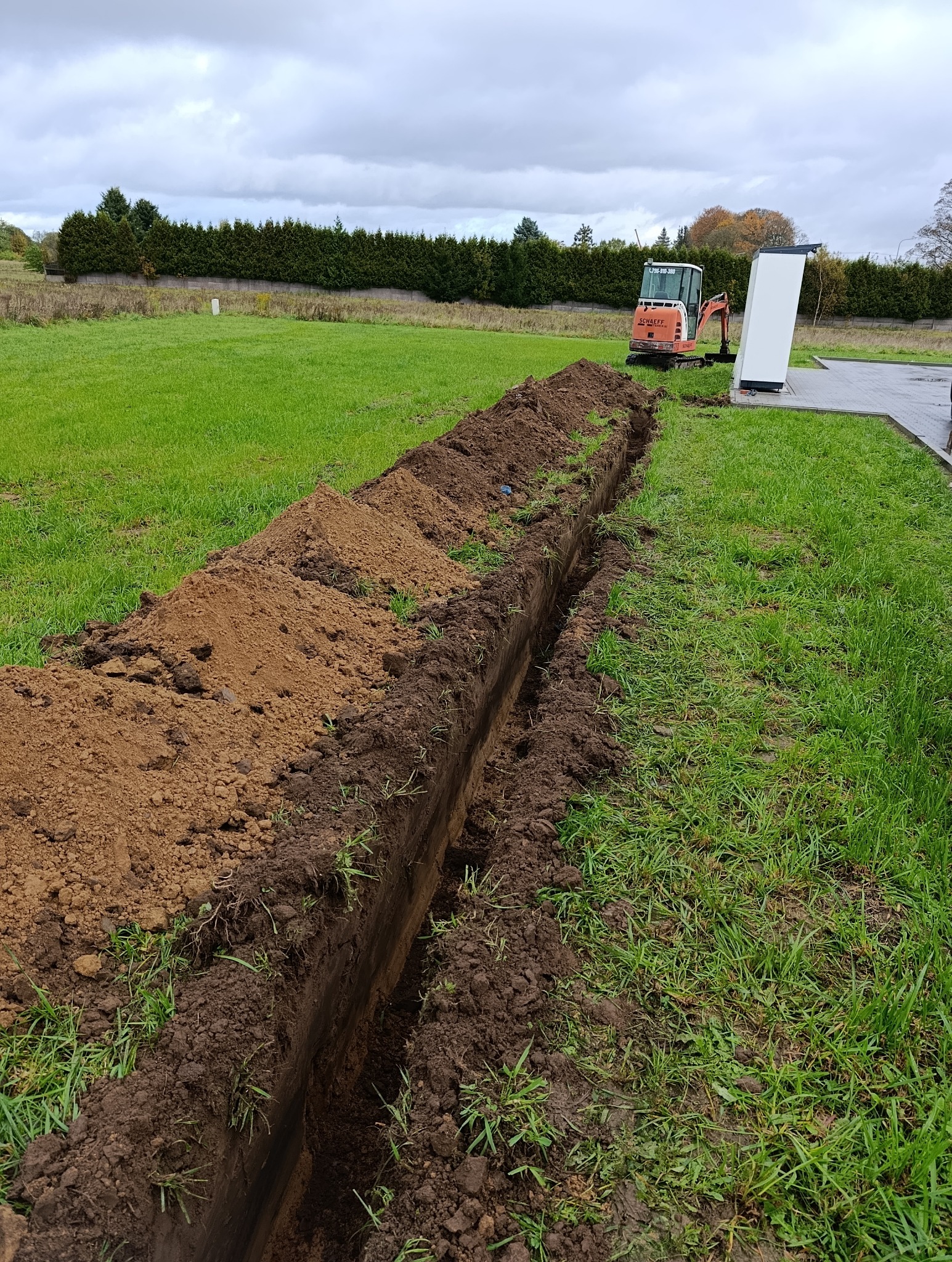 Wykopany rów w trawie, widoczny mały pomarańczowy traktor. Świeżo wykopana ziemia leży obok rowu. W tle żywopłot i pochmurne niebo.