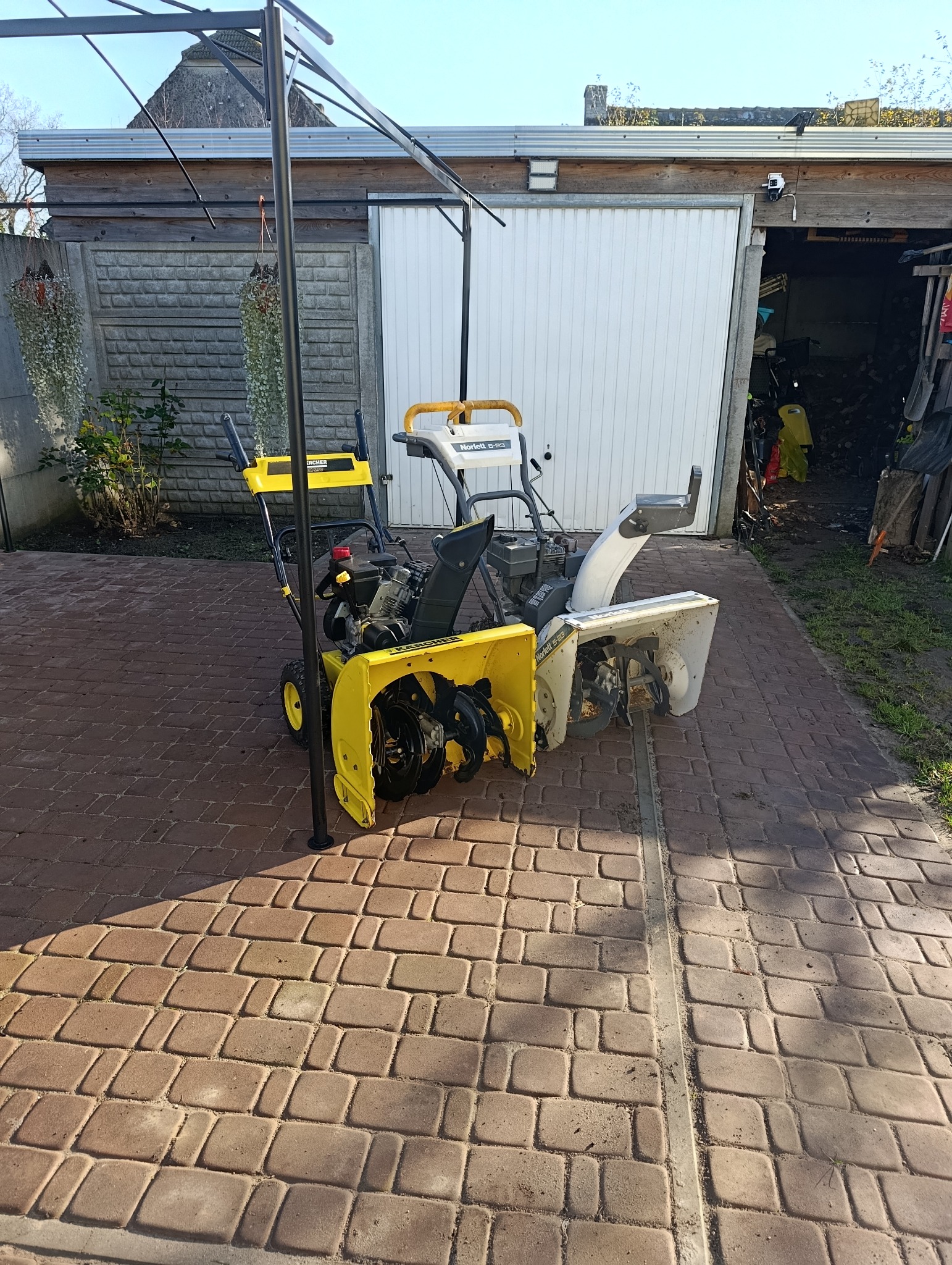 Dwie odśnieżarki (żółta i biała) stoją na kostce brukowej przed garażem. Widoczne elementy konstrukcyjne zadaszenia. Słoneczny dzień.