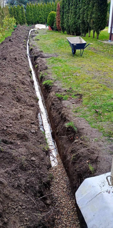 Długi wykop w ziemi z ułożoną białą rurą drenażową, częściowo zasypaną żwirem. W tle trawnik, drzewa iglaste i taczka. System drenażowy w ogrodzie.