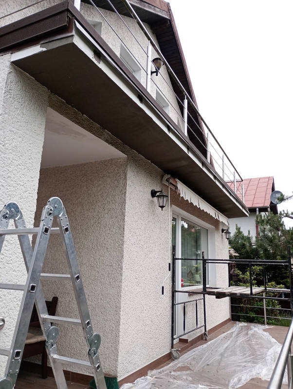Elewacja domu po renowacji z rusztowaniem i drabiną. Jasna fasada z balkonem i metalową balustradą. Widoczne lampy zewnętrzne i okno.