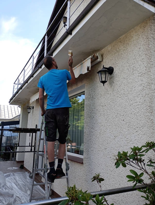 Mężczyzna na drabinie maluje elewację domu pędzlem. Widoczne zabezpieczone okno, lampa zewnętrzna i balkon z metalową barierką. Ujęcie z dołu.