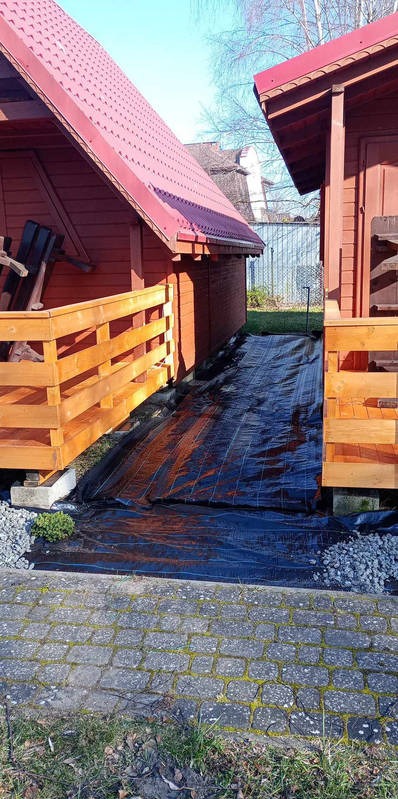 Przygotowanie podłoża pod trawnik z rolki: rozłożona czarna agrowłóknina między drewnianymi domkami, widoczne kamienie i kostka brukowa. Słoneczny dzień.