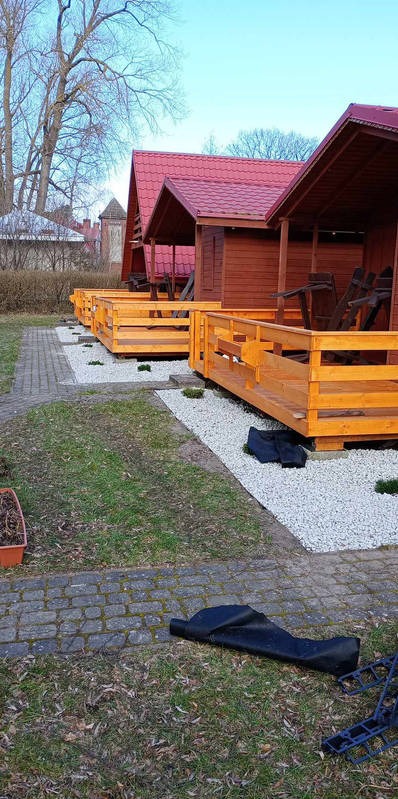 Rząd drewnianych domków letniskowych z czerwonymi dachami, oddzielonych od trawnika białym żwirem. Widoczne elementy konstrukcyjne i otoczenie.