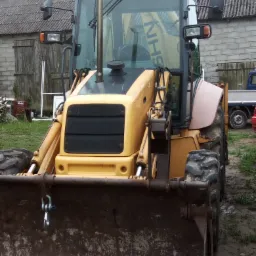 Żółta koparko-ładowarka z widoczną łyżką z przodu, stojąca na błotnistym terenie, z budynkiem gospodarczym w tle.