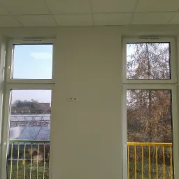 Dwa białe okna PCV w jasnym pomieszczeniu; przez jedno widać szklarnię, przez drugie drzewo bez liści i żółtą barierkę balkonu.