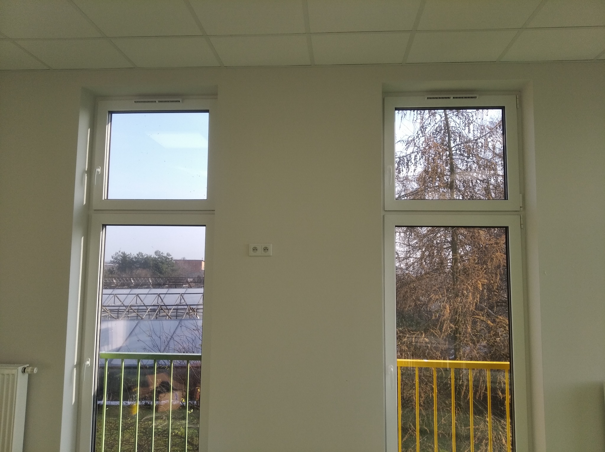 Dwa białe okna PCV w jasnym pomieszczeniu; przez jedno widać szklarnię, przez drugie drzewo bez liści i żółtą barierkę balkonu.