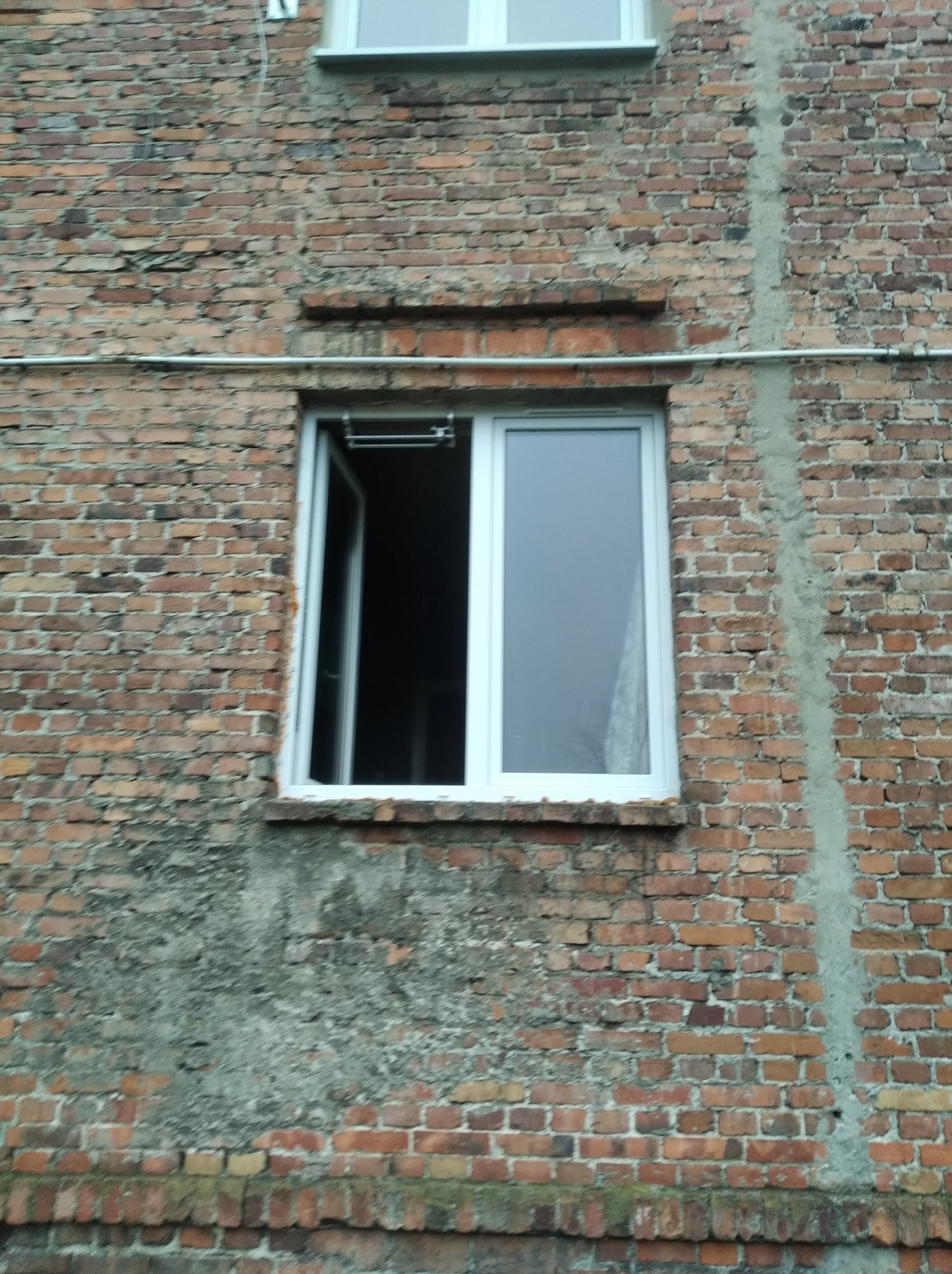 Białe okno PCV częściowo otwarte na tle starej ceglanej ściany budynku z widocznymi śladami napraw i zabrudzeń.